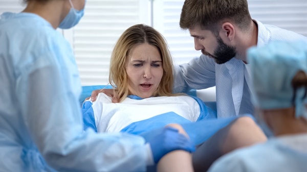 A distressed woman giving birth in a hospital bed, surrounded by medical staff in blue gowns while a supportive man leans over and touches her shoulder.